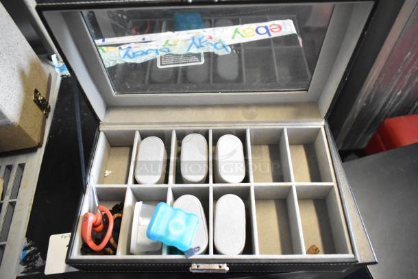 Black watch case with glass lid and multiple compartments, some filled with objects. Includes label reading "For N".