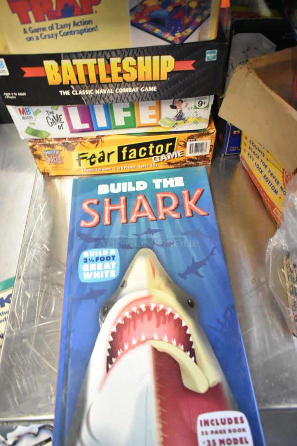 Tier lot of board games and books, featuring "Battleship," "The Game of Life," "Fear Factor," and "Build the Shark" kit.