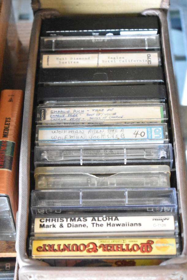 Collection of cassette tapes in a box, featuring various artists and titles. Includes labels and handwritten notes.
