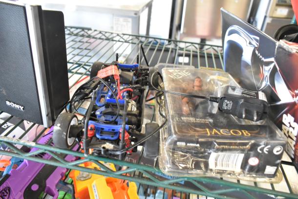 Two-tier shelf with a Sony speaker, Jacob doll in packaging, a Star Wars Darth Vader box, and colorful jerseys. Mixed items.