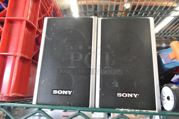 Sony speakers, black mesh with silver trim, placed on a shelf. Visible moderate wear; part of a mixed auction lot.