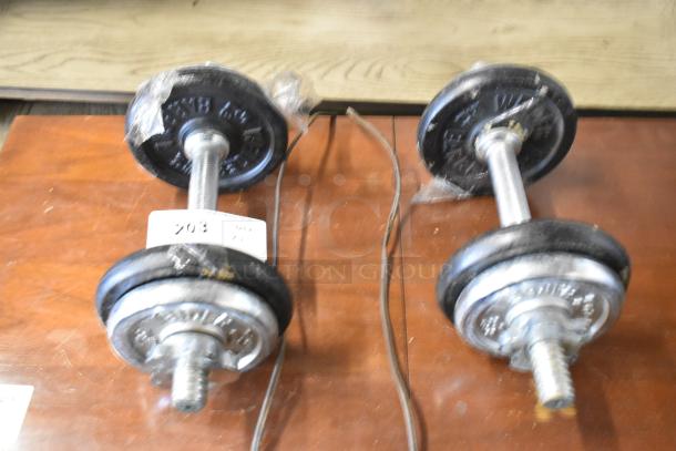 Two metal dumbbells on a wooden surface, labeled with auction tag 203. Each has adjustable weight plates.