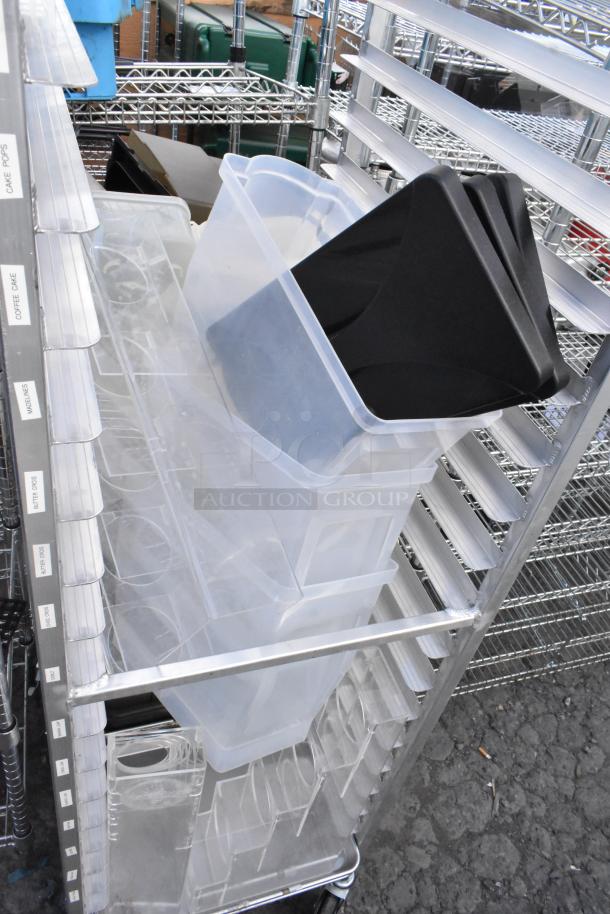 Metal commercial pan transport rack with contents, on commercial casters, featuring multiple clear plastic bins and black trays. Tags on the side indicate various pastries.