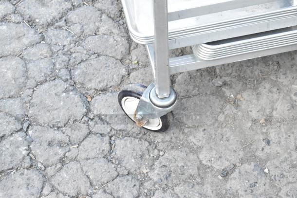 Metal commercial pan transport rack, sturdy metal frame, features commercial casters for mobility, visible on rough surface.