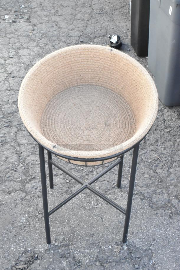Woven beige basket on metal stand, outdoor setting, textured ground surface. No visible tags or markings.