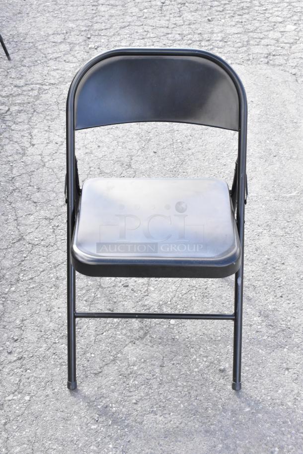 Black folding chair on outdoor pavement; metal frame, used condition. Part of auction set with cart and shelving unit.