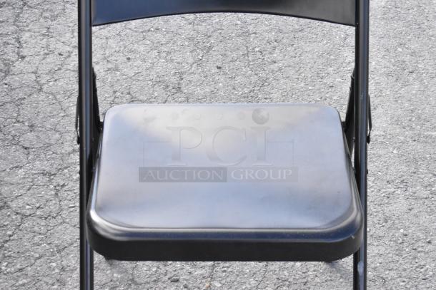 Black metal folding chair, slightly worn, set on asphalt. Part of auction lot with multiple items.