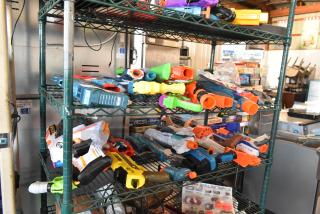 Two-tier shelf with assorted toy guns in varying colors. Includes blasters, dart guns, and accessories. Some packaging visible.