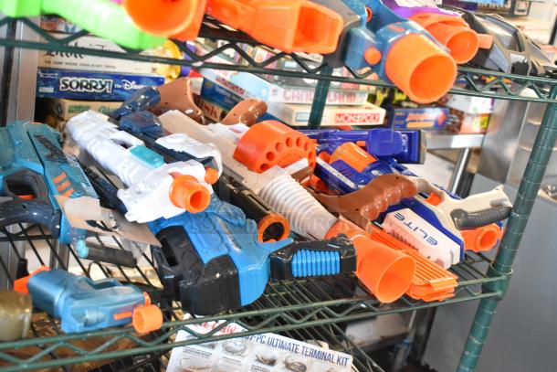 Two-tier shelf with various toy guns, including models with "Elite" branding. Mix of colors, featuring blue and orange.