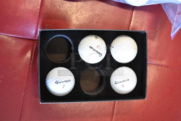 Lot of various golf balls in black box, featuring TaylorMade branded balls numbered 2, 3, and 5. Some show minor wear.