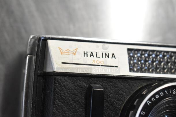 Halina 300 film camera detail, featuring leather texture and a metallic crown logo. In good condition.