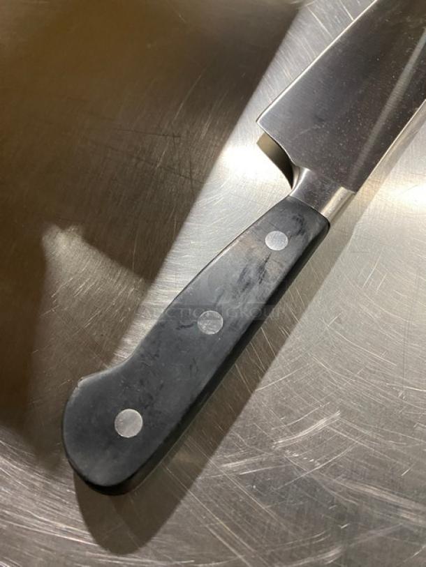 Commercial stainless steel chef knife with black grip handle, visible rivets, shown on metallic surface. Mint condition.