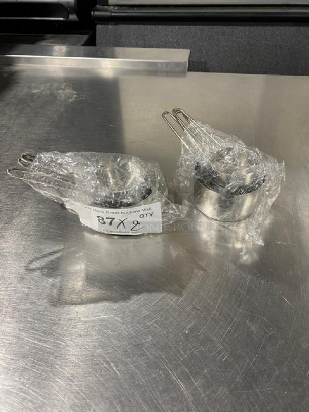 Set of new commercial stainless steel measuring cups, wrapped in plastic. Label shows "87 Qty 2."