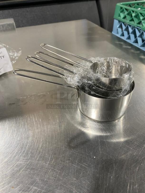 New commercial stainless steel measuring cup set, wrapped in plastic. Four nested cups with straight handles on a metal surface.