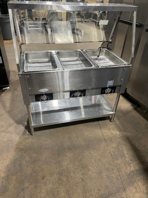 Serv-Ware stainless steel hot food table, model EST3-1, with 3 open wells, sneeze guard, electric-powered, on legs.