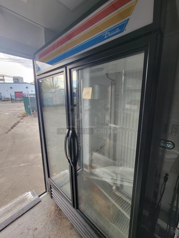 True Refrigeration GDM-43F-HC-TSL01 freezer with glass doors inside SDG trailer, showcasing wire shelving and visible outdoor scene.