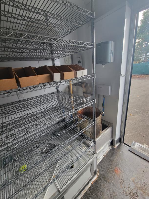 Interior of SDG 20' Goodees trailer with multi-tier chrome wire shelving, hand sanitizer dispenser, and floor drainage area visible.