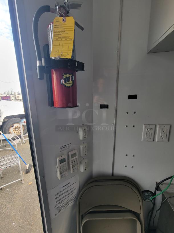 Red fire extinguisher mounted on a wall inside the SDG trailer with a yellow inspection tag. Visible electrical outlets and climate control units nearby.