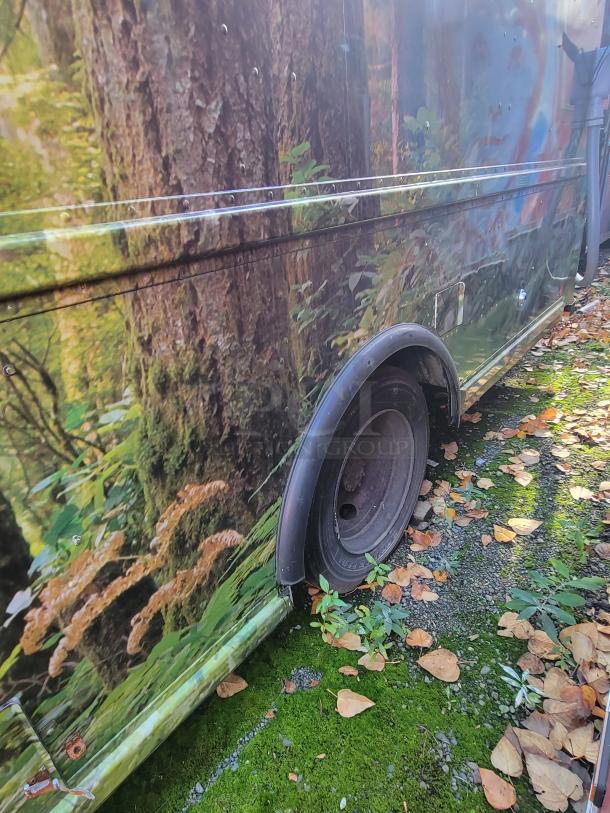 2004 Freightliner MT45 mobile kitchen side view. Forest-themed wrap, visible fender, autumn leaves, and mossy ground.