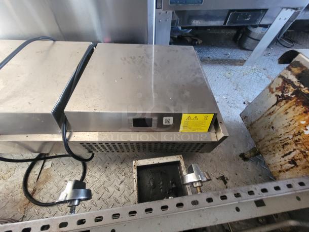 Stainless steel commercial-grade appliance in mobile kitchen trailer on diamond plate floor. Features warning labels and visible power cords.