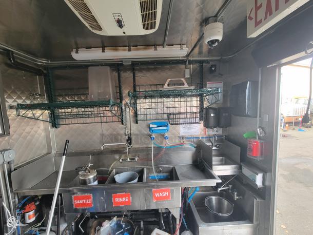 Interior of a 2022 Titanium 28' Food Trailer featuring stainless steel sinks labeled "Sanitize," "Rinse," and "Wash." Includes overhead storage racks, a handwashing station, and installed fire safety system, ideal for high-volume food service operations.
