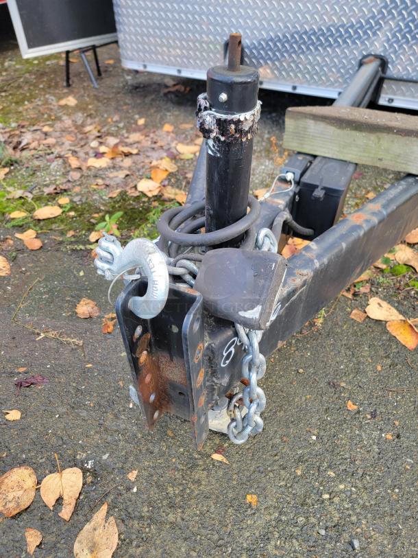 Trailer hitch of 2021 24’ Diamond Cargo Mobile Kitchen, showing rust, safety chain, and coiled cable on gravel surface.