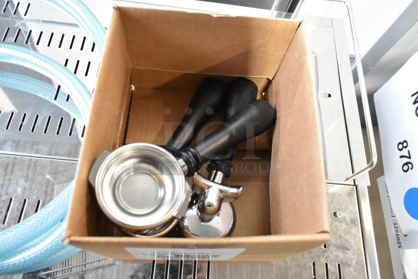 Box containing three portafilters with black handles for Estella ECEM2 espresso machine. Visible blue hoses nearby.