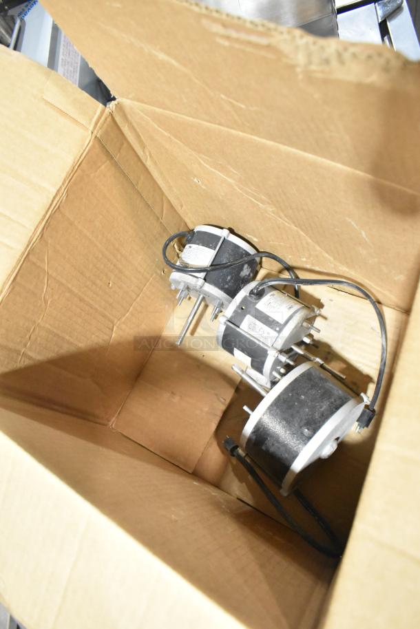 Three various motors in a cardboard box, each with visible labels and wiring. Slight wear evident.