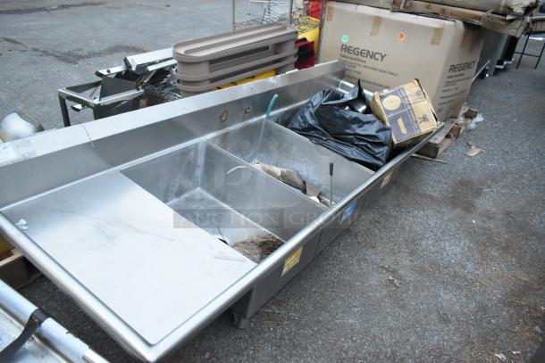 Stainless steel 3 bay sink with dual drain boards, no legs, visible wear, includes various labels and packaging nearby.