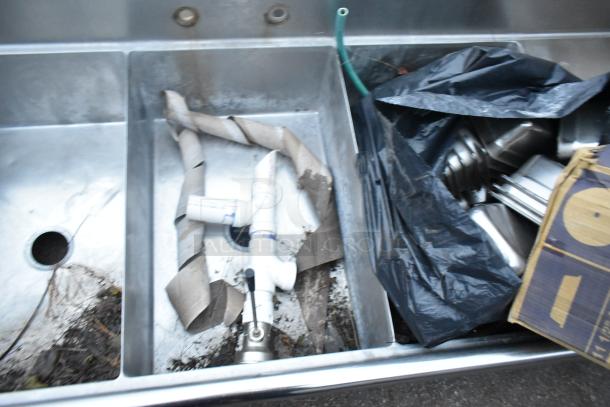 Stainless Steel 3 Bay Sink with Dual Drain Boards, no legs, visible debris and plumbing parts, in used condition.