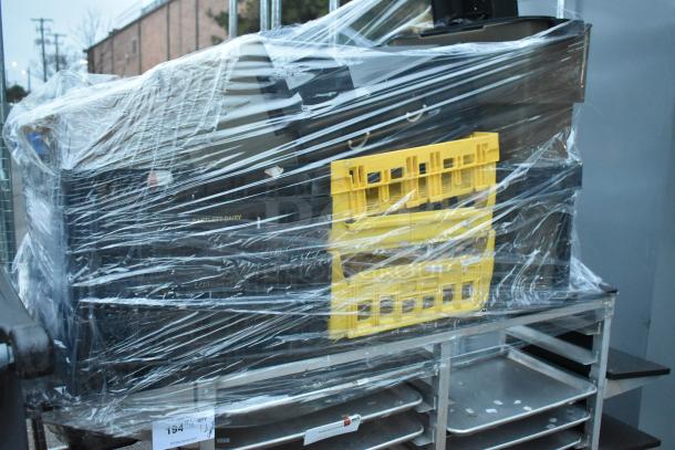 Metal commercial double pan transport rack on commercial casters, wrapped with plastic, contains trays and yellow crates.