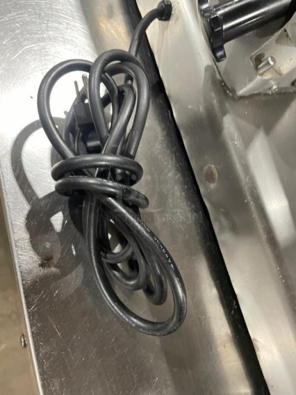 Stainless steel deli meat slicer with power cord on countertop. Heavy-duty commercial model, shows wear.