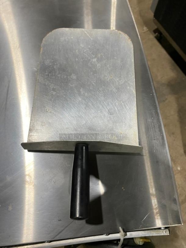 Metal pizza peel with a black handle on a stainless steel surface. Scratch marks visible.