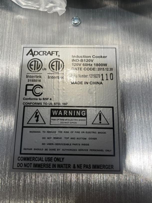 Alt text: "Adcraft INDB120V induction cooker, stainless steel, model 12150270, new condition. Label shows electrical and safety info."