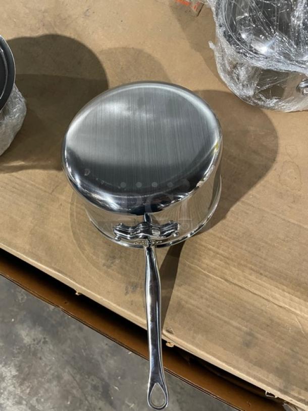 Brand new Mauviel M'Cook stainless steel 5-ply saucepan, includes lid and cast handle, shown on a cardboard surface.