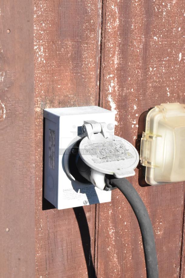 Outdoor electrical hookup with a plugged-in cable on a weathered wooden wall.