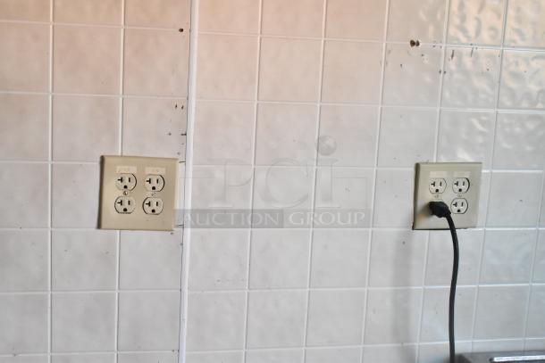 Food trailer interior with white tiled walls and two outlet panels. One outlet has a plugged-in black cord.