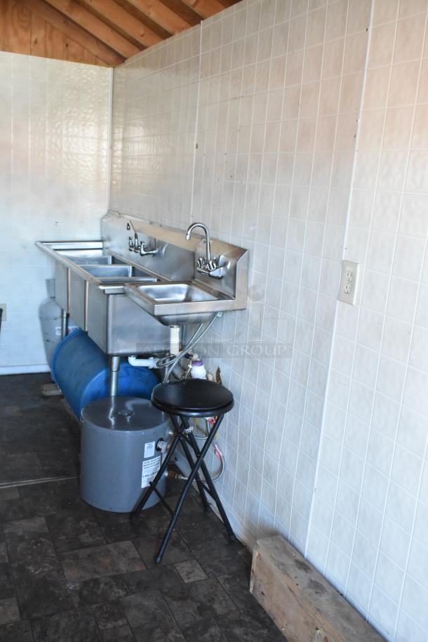 Food trailer interior with stainless steel triple sink setup, foldable stool, blue storage barrel, and white tiled walls.