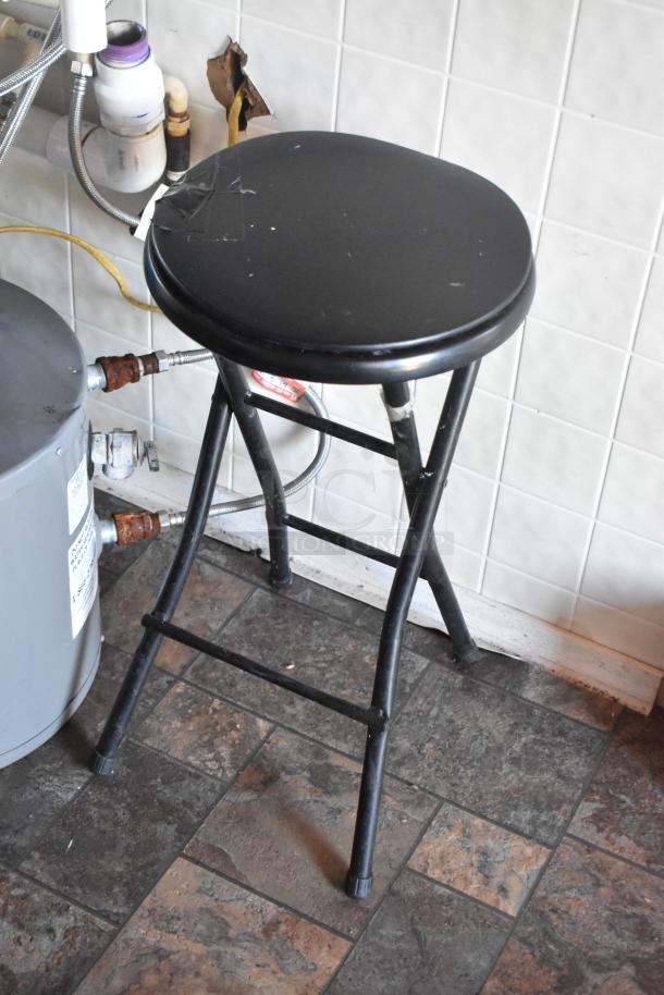 Black metal stool with a padded seat, minor wear visible, placed on a tiled floor, pipes visible in the background.