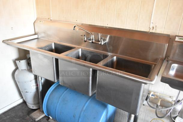 Stainless steel three-compartment sink inside a food trailer, equipped with faucet, propane tank, and blue barrel underneath.