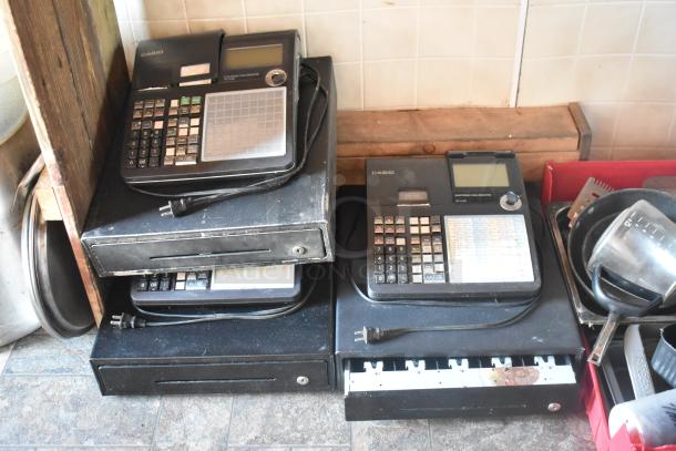 Stacked black Casio electronic cash registers with attached power cords, resting on cash drawers.