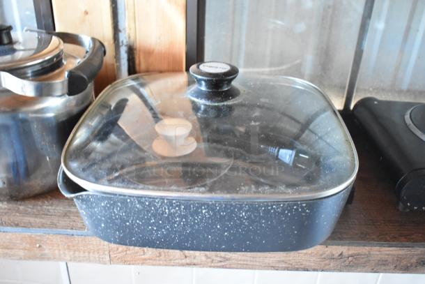 Enamel cooking pan with glass lid, on food trailer counter. Visible plug inside pan. Ideal for mobile kitchen use.