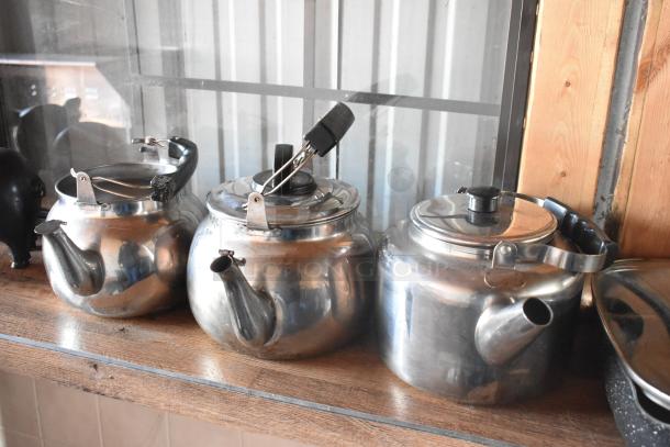 Three stainless steel kettles with black handles and spouts, displayed on a wooden shelf in a food trailer.
