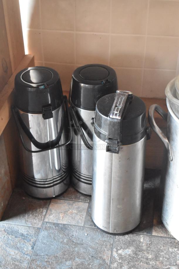 Three stainless steel beverage dispensers with black lids and handles, placed on a tiled floor.