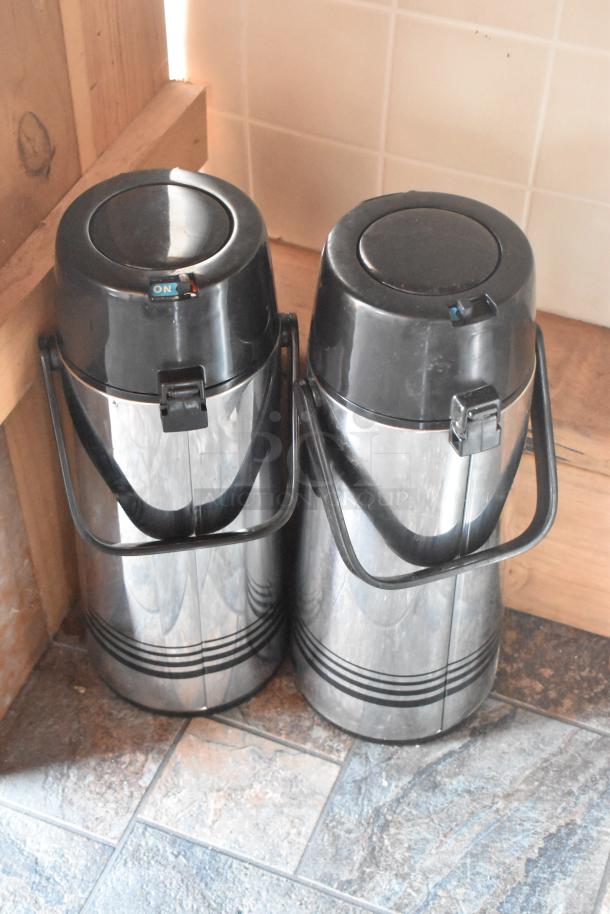Two stainless steel thermal beverage dispensers with black lids and handles, labeled with "ON" switches, on tile floor.