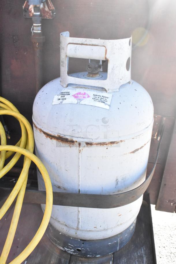 White propane tank secured with a metal strap, showing wear. Label indicates "Liquefied Petroleum Gas, UN 1075." Yellow hose visible nearby.