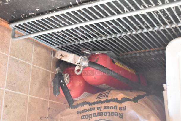 Red fire extinguishers stored under a metal shelf in a tiled area of the 2007 Lanchester food trailer.