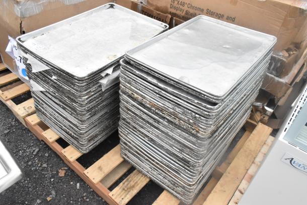 106 metal full-size baking pans stacked on pallets, showing wear and use. Tags visible indicating auction quantity.