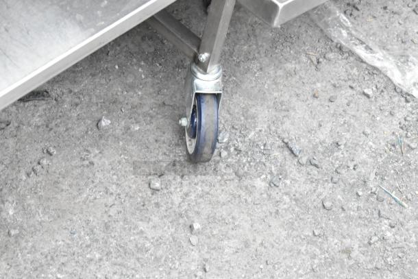 Stainless steel table wheel detail, showing blue commercial caster in good condition on a rough concrete surface.