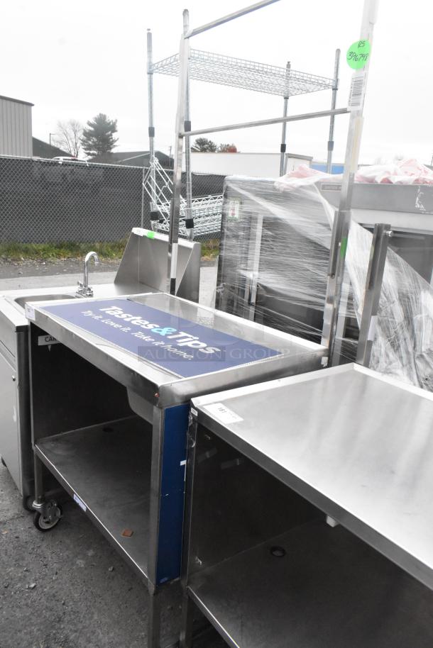Stainless steel table with under shelf, drawer, and commercial casters. Features integrated sink and branded surface. Visible tags indicate auction ID.